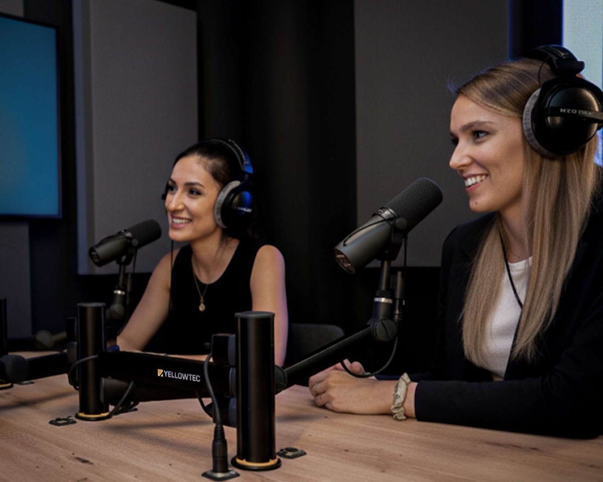 Zwei Moderatorinnen mit Kopfhörern sprechen am Studiotisch in Broadcast-Mikrofone